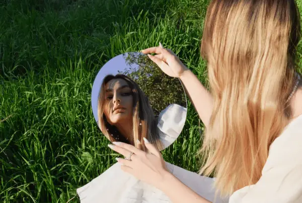 Mulher sentada na grama, olhando o próprio reflexo em um espelho redondo