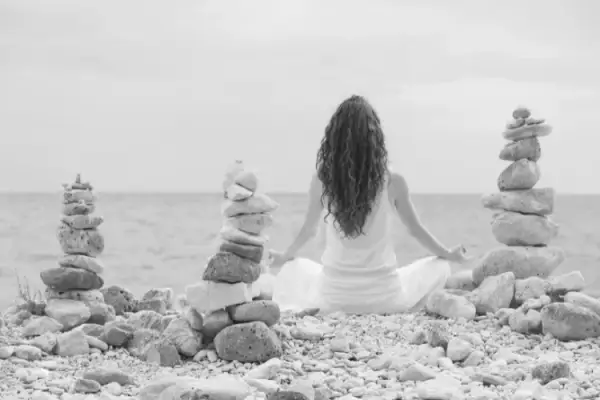 Mulher meditando de costas na praia, entre pilhas de pedras