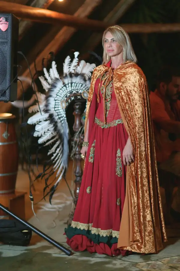 Valéria Tavares de vestido vermelho e capa dourada, ao lado de um cocar de penas