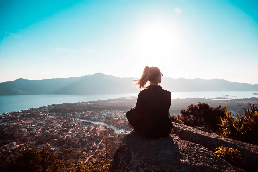 Mulher sentada sozinha no alto de uma pedra, contemplando o horizonte ao pôr do sol