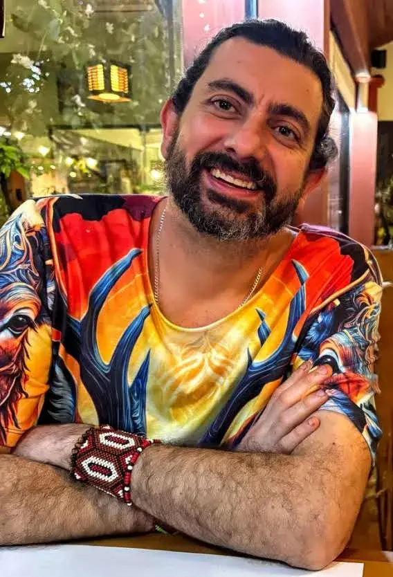 Rodrigo Otávio sorrindo, de braços cruzados, com camiseta colorida e pulseira indígena